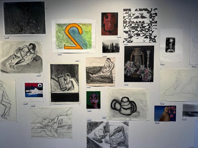 collection of student art on gallery wall. My favorite is a colored charcoal drawing of a lamp with a candlestick and roses around it. It feels bleak and drained of life. Although it is in color, the colors are muted for effect.